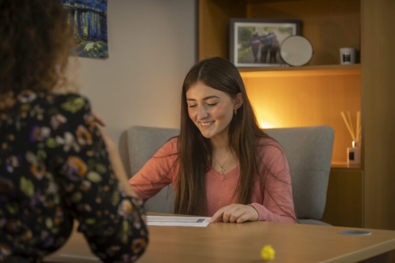 A young woman smiling while talking with someone across a table in a warmly lit room, papers on the table between them.