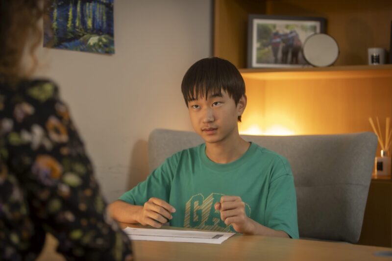 Student in a green t-shirt sitting at a table, focused on writing, with another person visible in the foreground.