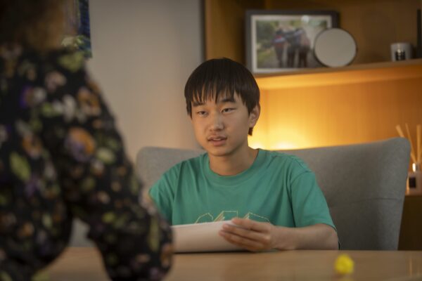 A student sitting on a sofa holding a sheet of paper, talking with someone across a table in a warmly lit room.