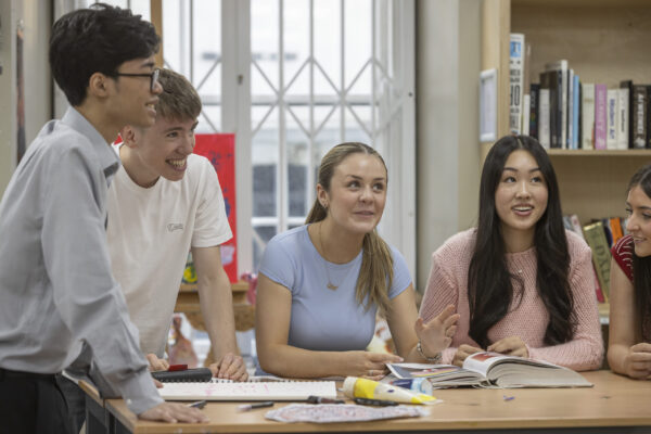 Four young adults sit and stand around a table in a library or classroom, smiling and talking, with books and papers in front of them. Shelves with books and other items are visible in the background.
