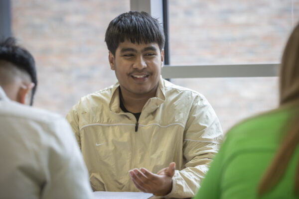 A student smiling while talking with classmates seated around a table.