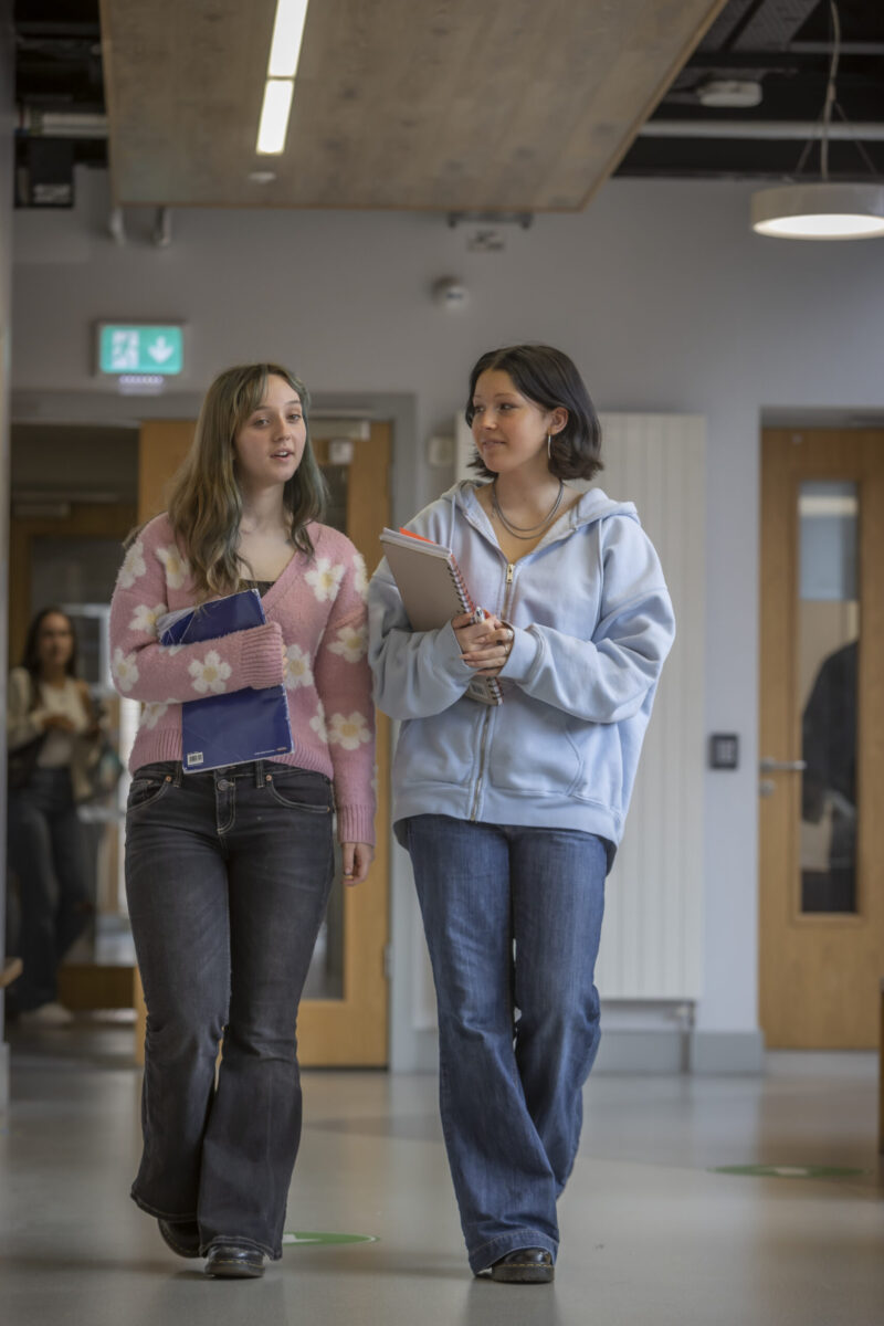 two students walk in a brightly lit hallway, chatting casually