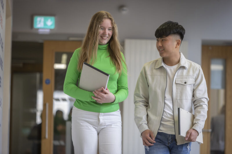 Two students smiling and chatting as they walk together down a corridor, carrying folders.