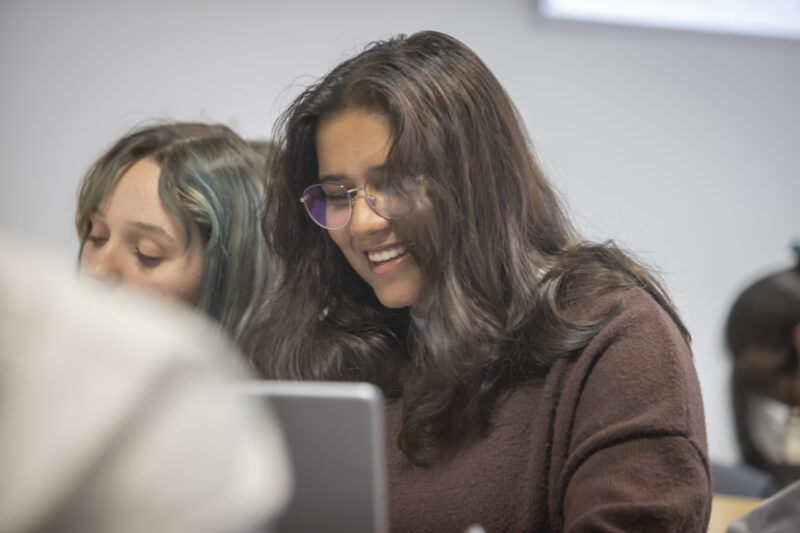 A student smiling while looking at a laptop, with classmates beside her.