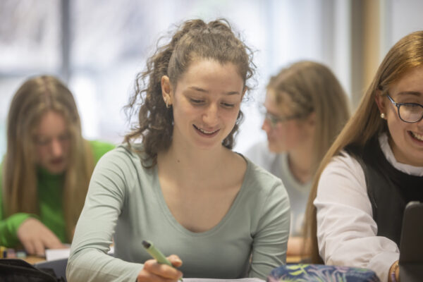 A student writing at a desk during class, with classmates visible behind her.