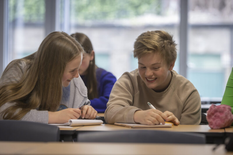 Three students smiling and working together at a desk, writing in notebooks during class.