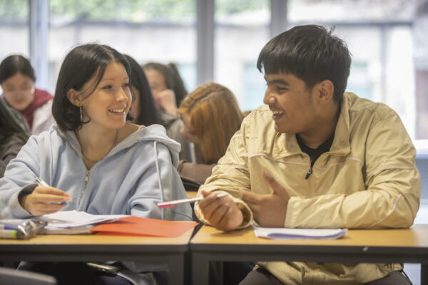 Two students sit at a desk in a classroom, smiling and talking to each other whilst holding pens and taking notes, with other students visible in the background.