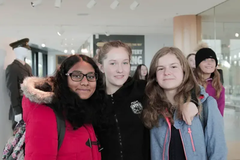 Three students smiling together indoors, with others visible in the background.