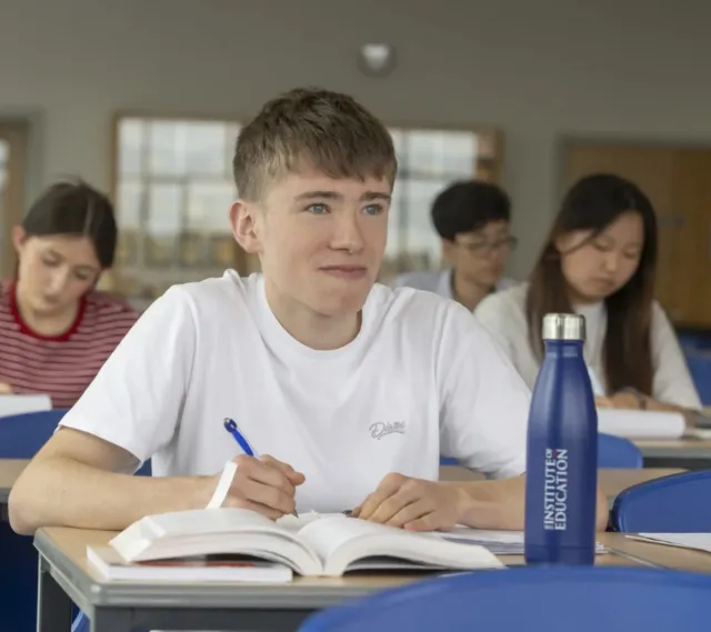 A student in a white shirt writes in a notebook at a desk with an Institute of Education water bottle. Other students study in the background in a classroom.