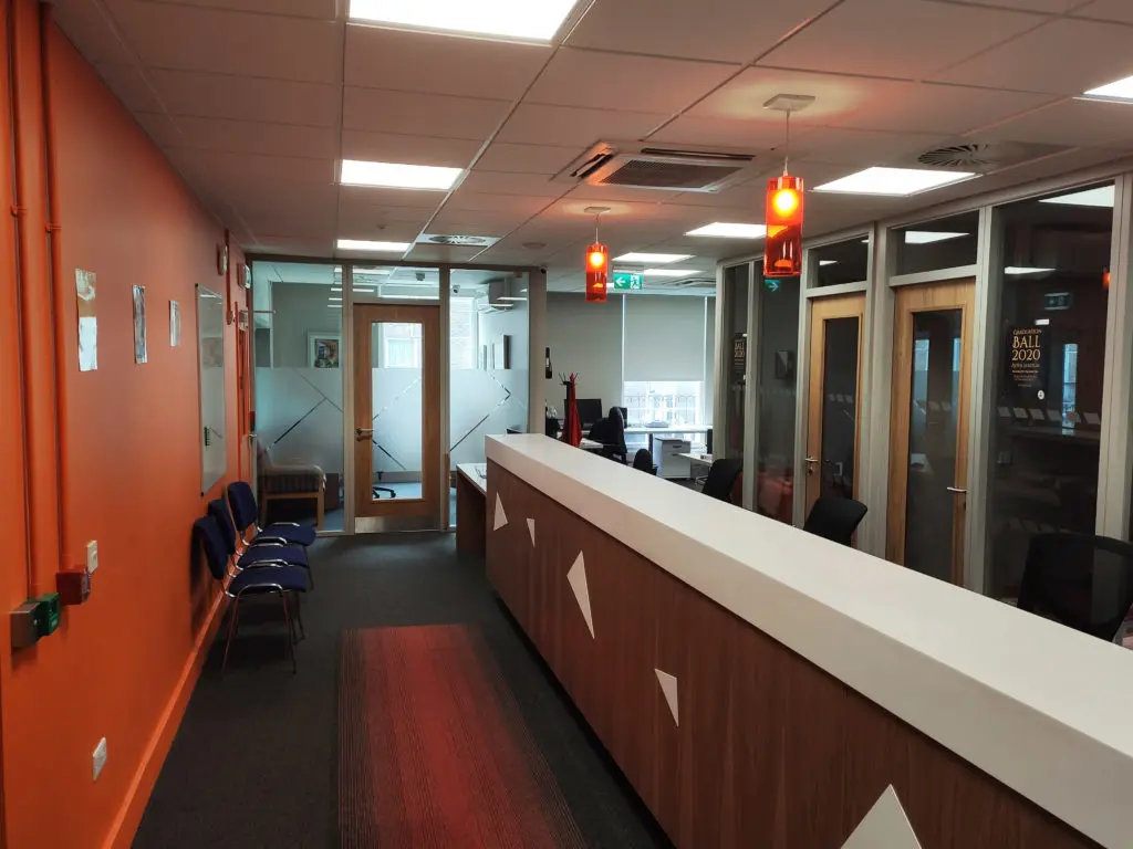 A reception desk area with an orange feature wall, waiting chairs, and glass-partitioned offices beyond.
