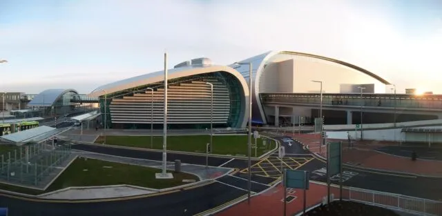 Modern curved glass and steel building, resembling Dublin Airport's terminal, with roads and signage in the foreground.
