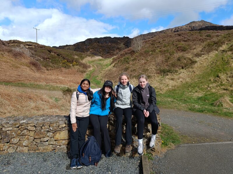 Four students posing together on a hiking trail, with hills and cloudy sky in the background.