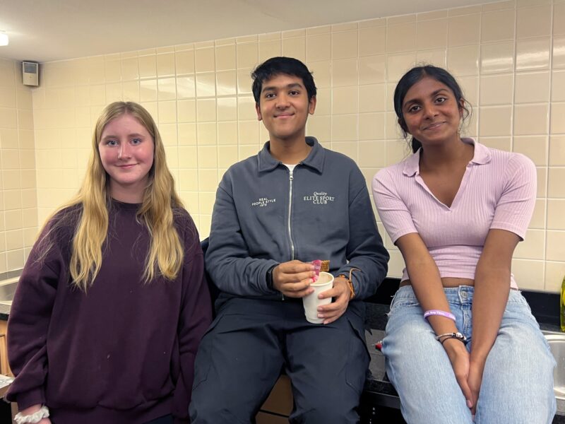 Three students smiling together, one holding a takeaway drink, in a tiled kitchen or canteen area.
