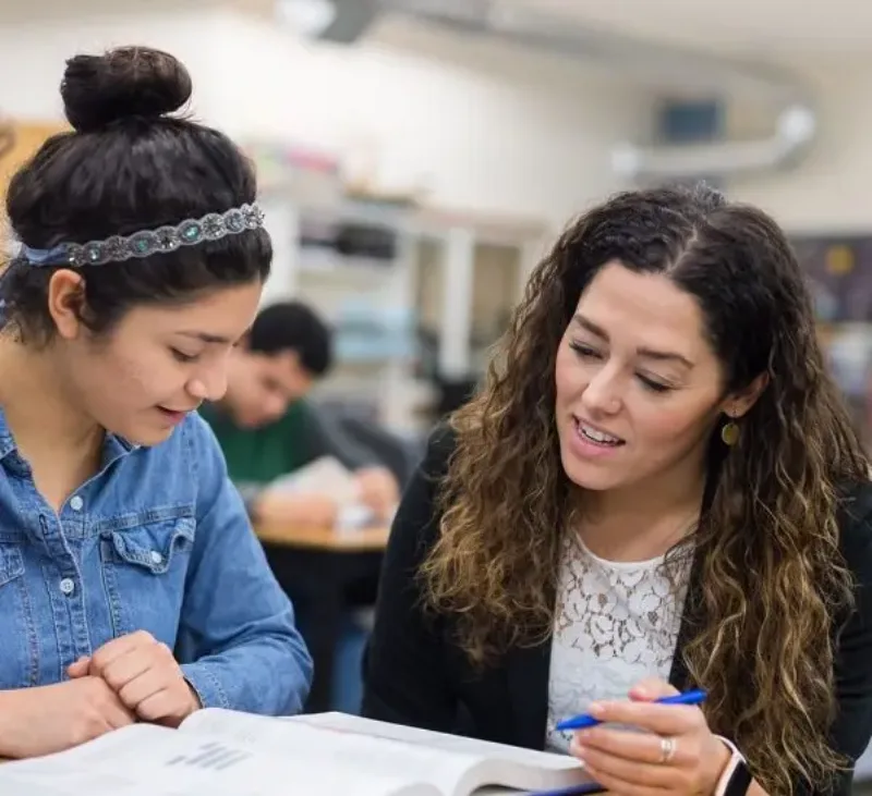 A teacher helping a student with schoolwork at a desk, pointing at a page with a pen.