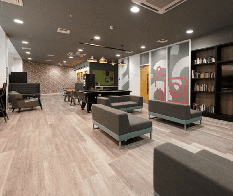 Interior of a student common room at Binary Hub, with grey sofas, wooden flooring, a breakfast bar, bookshelves and warm pendant lighting.