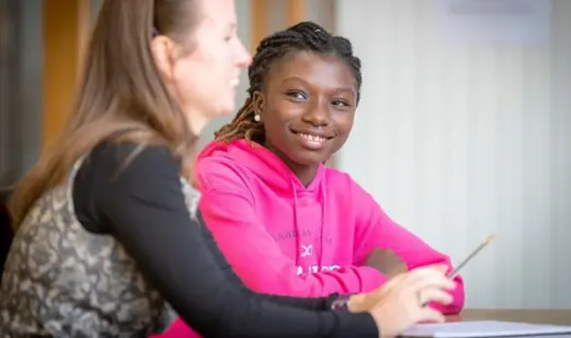 A smiling student in a pink hoodie sitting at a table with a teacher, looking towards the camera.