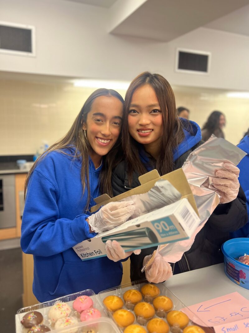 Two students in blue jumpers and gloves smiling while holding baked goods and packaging, at a bake sale table with cakes on display.
