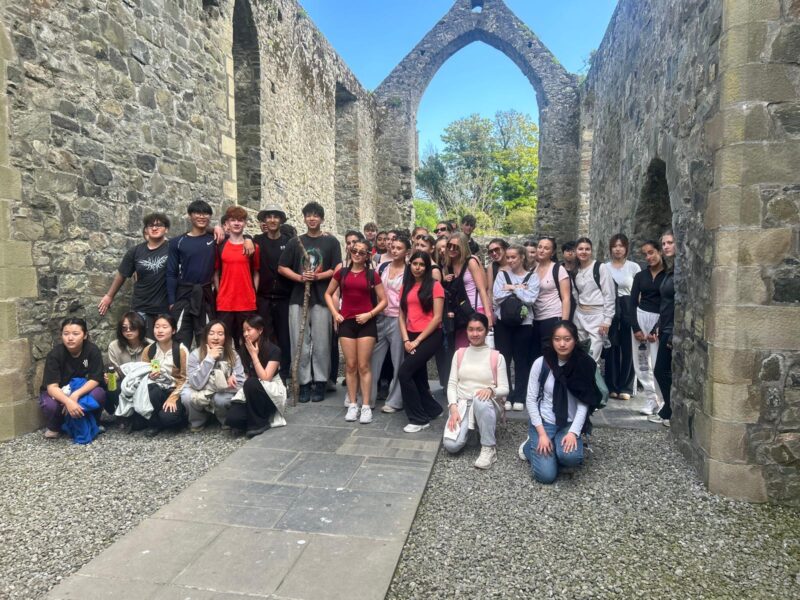 A large group of students posing for a photo inside the stone ruins of a roofless medieval church, on a school trip.