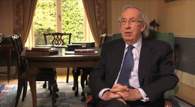 Raymond Kearns in a suit sitting in a wood-panelled study, being interviewed, with a window and antique furniture in the background.