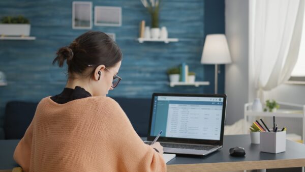 A student wrapped in a blanket, writing notes at a desk with a laptop showing a spreadsheet, in a home study setting.