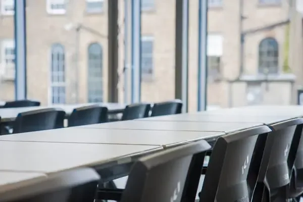 A close-up of a long table with black chairs in a meeting or study room, with tall windows showing brick buildings outside.