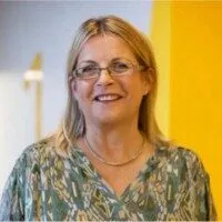 Smiling woman with blonde hair and glasses, wearing a green patterned blouse and necklace, stands in front of a bright yellow and white background, embodying the vibrant spirit of student life at the Institute of Education.