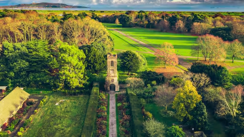 A stone tower stands amid lush gardens and pathways, surrounded by colourful trees and expansive green fields under a partly cloudy sky, with distant hills visible on the horizon—an inspiring setting reminiscent of the tranquil atmosphere found while attending leaving cert grinds in Killester.