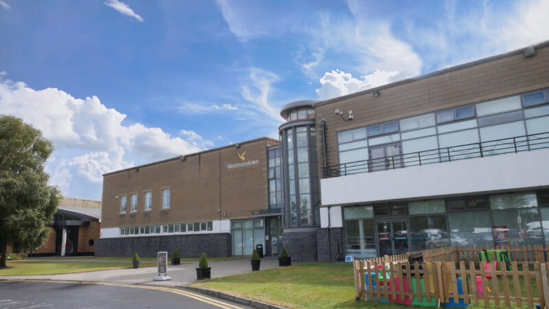 Modern building with large windows, a glass tower entrance, and a fenced play area in front—an inviting setting ideal for leaving cert grinds Westmanstown. The sky is bright with scattered clouds, and the building is bordered by a driveway and green lawn.