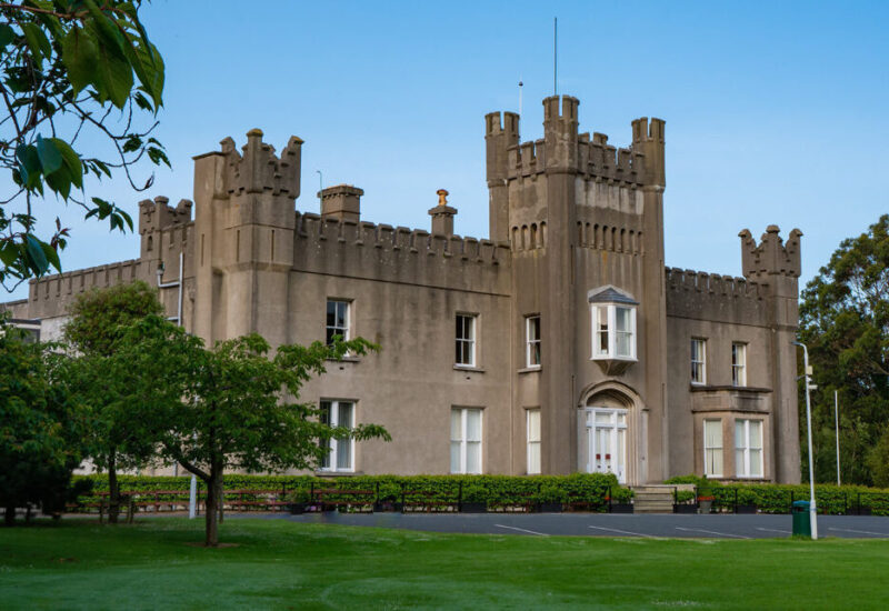 A historic grey stone building with castle-like battlements, arched windows, and a central entrance—offering Leaving Cert grinds Dalkey students can count on—stands surrounded by green lawns and leafy trees under a clear blue sky.