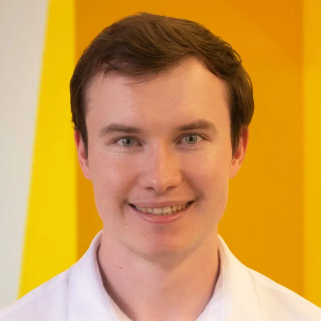 A young man with short brown hair smiles at the camera, wearing a white collared shirt. The background is yellow with geometric shapes.