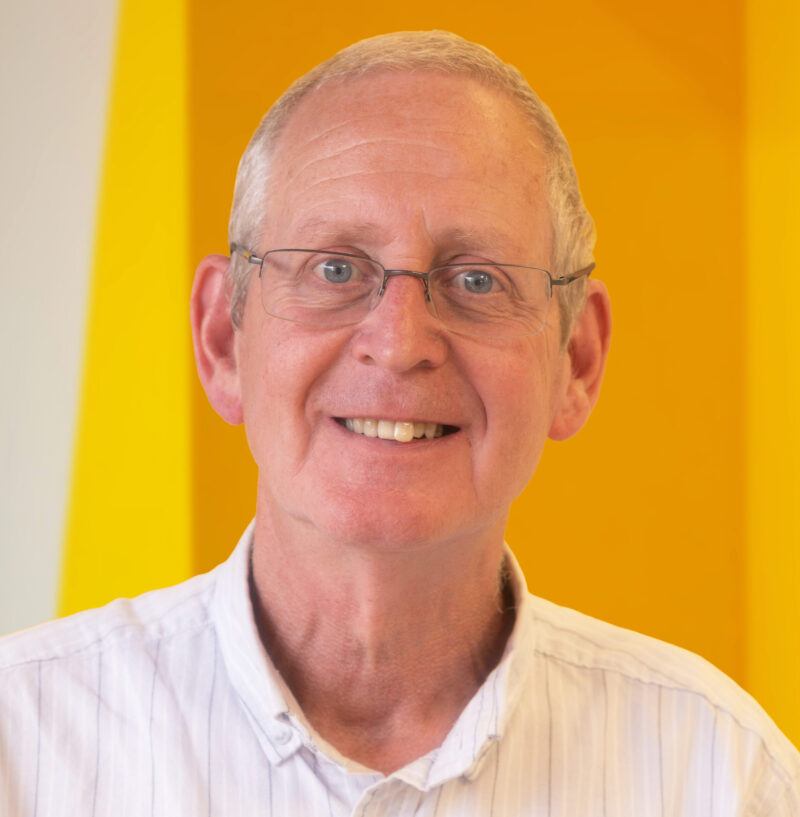 An older man with short grey hair and glasses smiles whilst wearing a light-coloured shirt. The background is bright yellow.