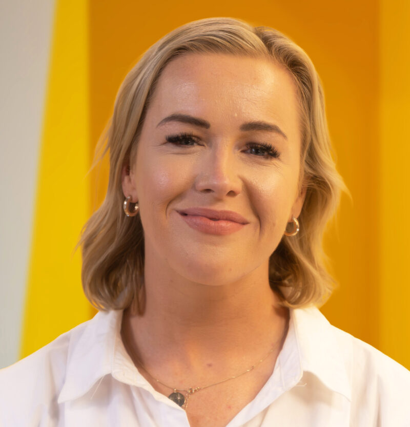 A woman with short blonde hair and hoop earrings smiles softly, wearing a white shirt and necklace, standing in front of a bright yellow and white background.