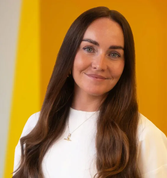 A woman with long brown hair and blue eyes, wearing a white top and necklace, smiles softly in front of a bright yellow background.