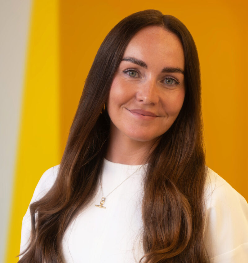 A woman with long brown hair and blue eyes, wearing a white top and necklace, smiles softly in front of a bright yellow background.