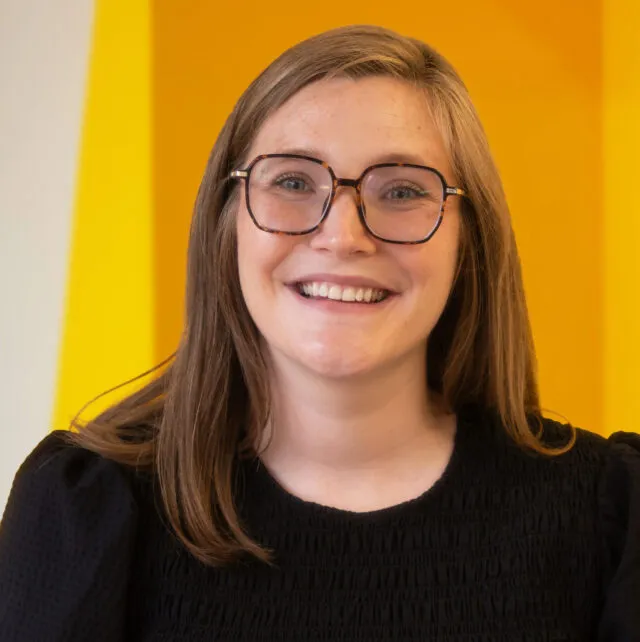 A woman with long brown hair and glasses smiles at the camera, wearing a black top and standing in front of a bright yellow background.