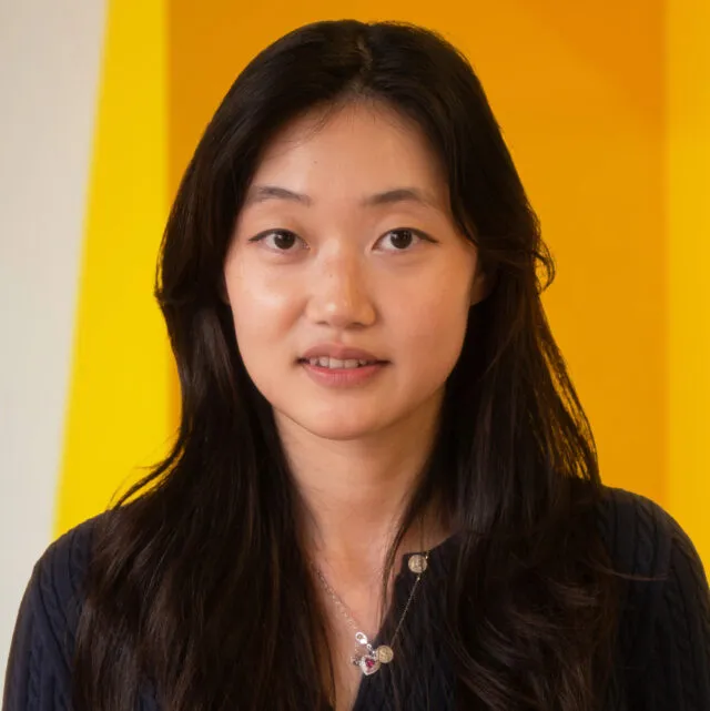 A woman with long dark hair and a navy top stands in front of a vibrant yellow background, looking at the camera and wearing a silver pendant necklace.