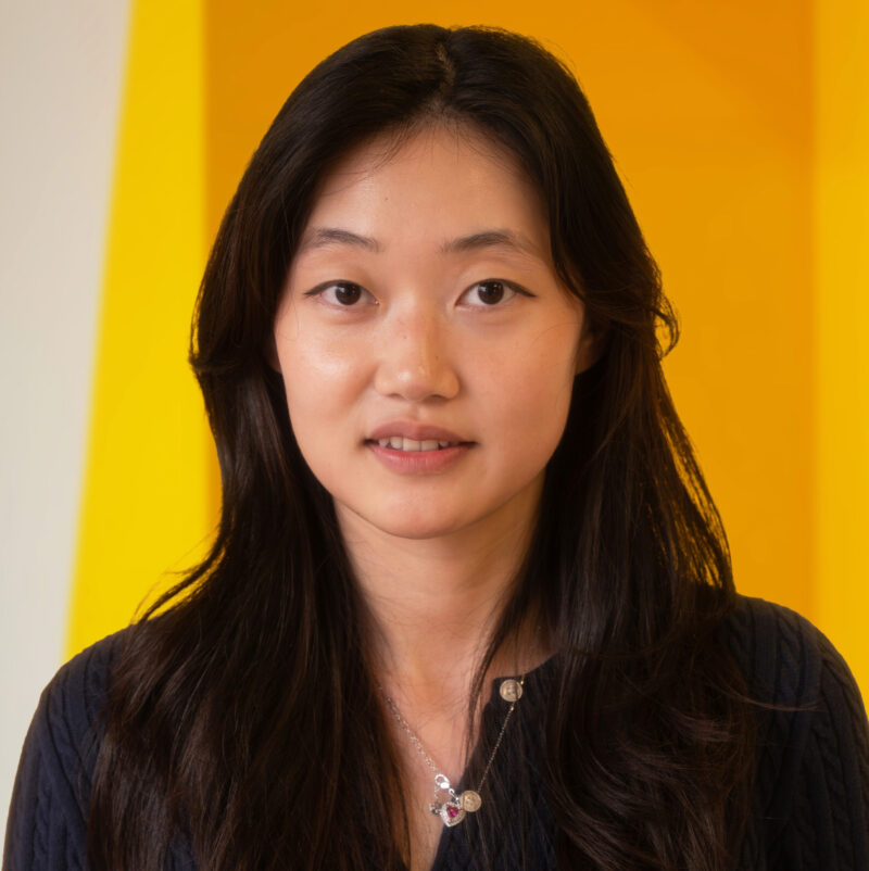 A woman with long dark hair and a navy top stands in front of a vibrant yellow background, looking at the camera and wearing a silver pendant necklace.