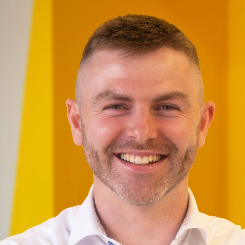 A smiling man with short hair and a trimmed beard, wearing a white collared shirt, stands in front of a bright yellow background.