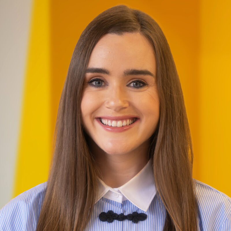 A woman with long brown hair, wearing a blue and white collared shirt, smiles at the camera. The background features bright yellow panels.