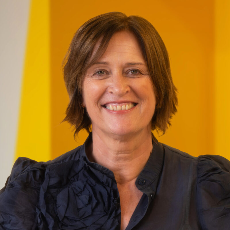A woman with short brown hair smiles at the camera, wearing a dark blouse with a frill on the shoulder, with a yellow background behind her.