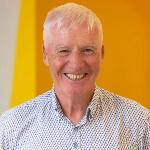 Older man with short white hair smiles, wearing a patterned button-up shirt. The background is bright yellow with a white stripe on the left side.