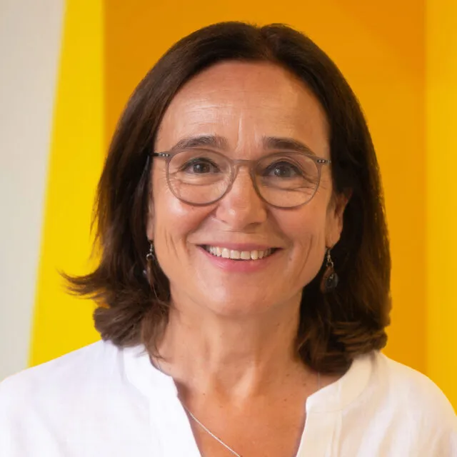 A woman with shoulder-length brown hair, glasses, and earrings smiles at the camera. She is wearing a white top and standing in front of a yellow background.