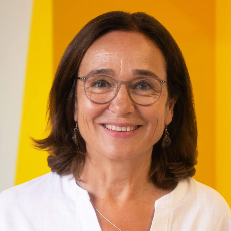 A woman with shoulder-length brown hair, glasses, and earrings smiles at the camera. She is wearing a white top and standing in front of a yellow background.