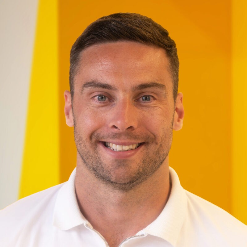 A man with short brown hair and a trimmed beard smiles at the camera, wearing a white collared shirt, standing in front of a bright yellow background.