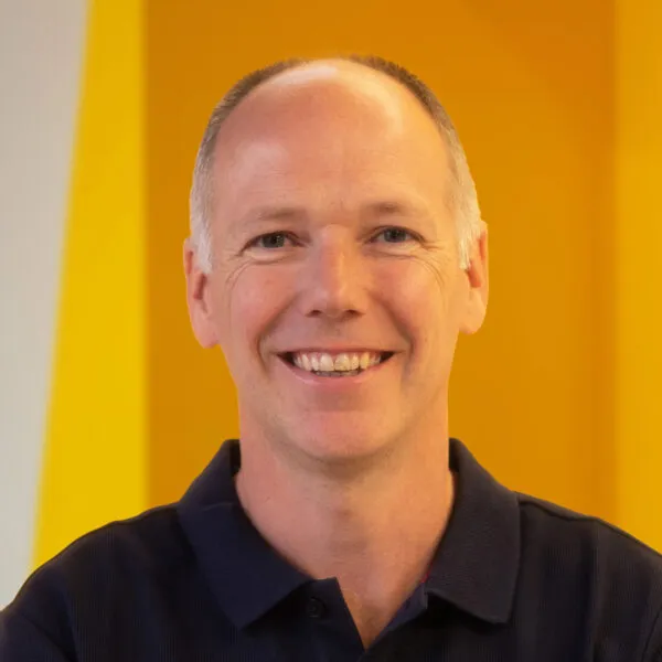A smiling person with short hair wearing a dark polo shirt stands in front of a bright yellow and orange background.