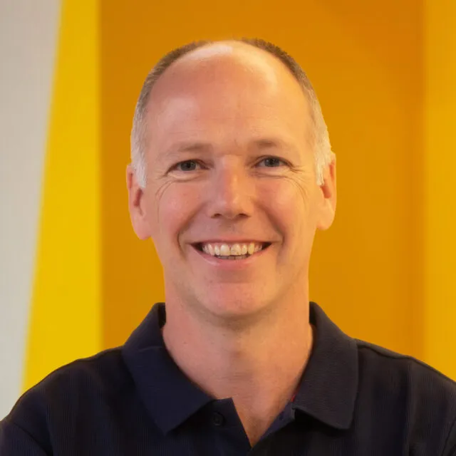 A smiling person with short hair wearing a dark polo shirt stands in front of a bright yellow and orange background.