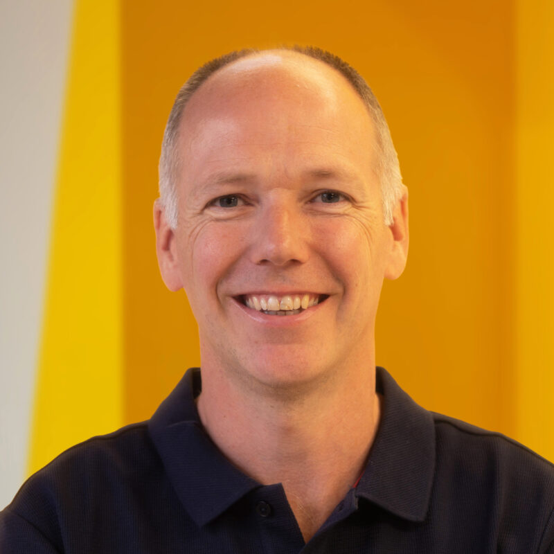 A smiling person with short hair wearing a dark polo shirt stands in front of a bright yellow and orange background.