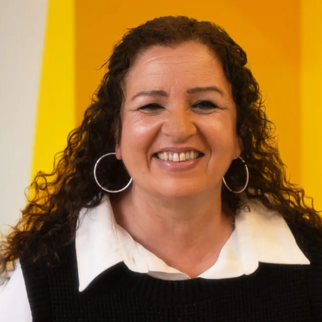 A woman with curly brown hair, large hoop earrings, and a white collared shirt under a black jumper smiles in front of a bright yellow background.