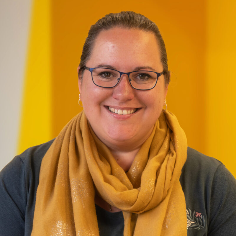 A woman with glasses and a yellow scarf smiles in front of a yellow background.