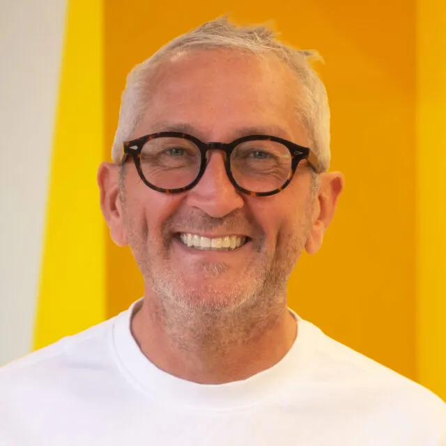 Smiling older man with short grey hair, wearing black glasses and a white T-shirt, stands in front of a bright yellow and white background.